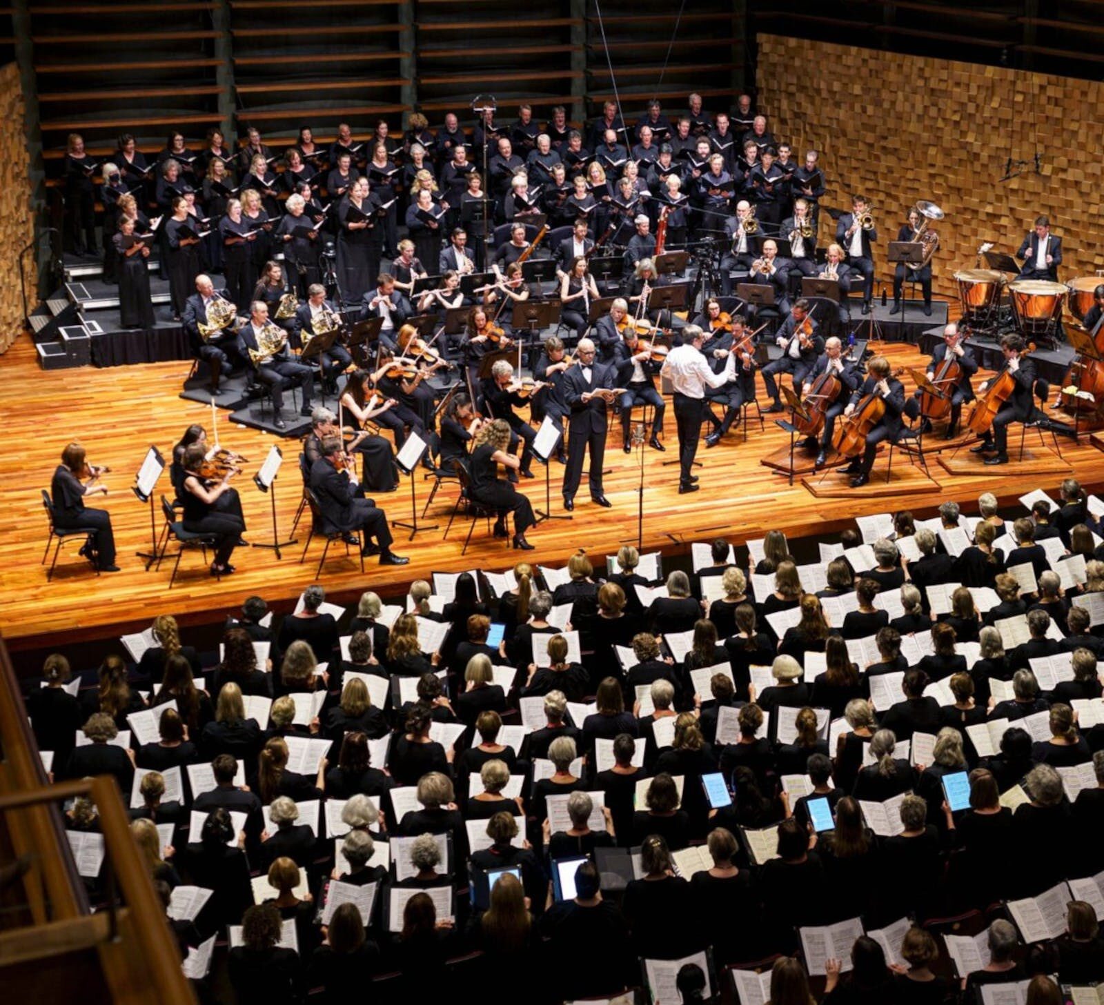 Photograph of a symphony orchestra. Choristers are situated behind and in front of them.
