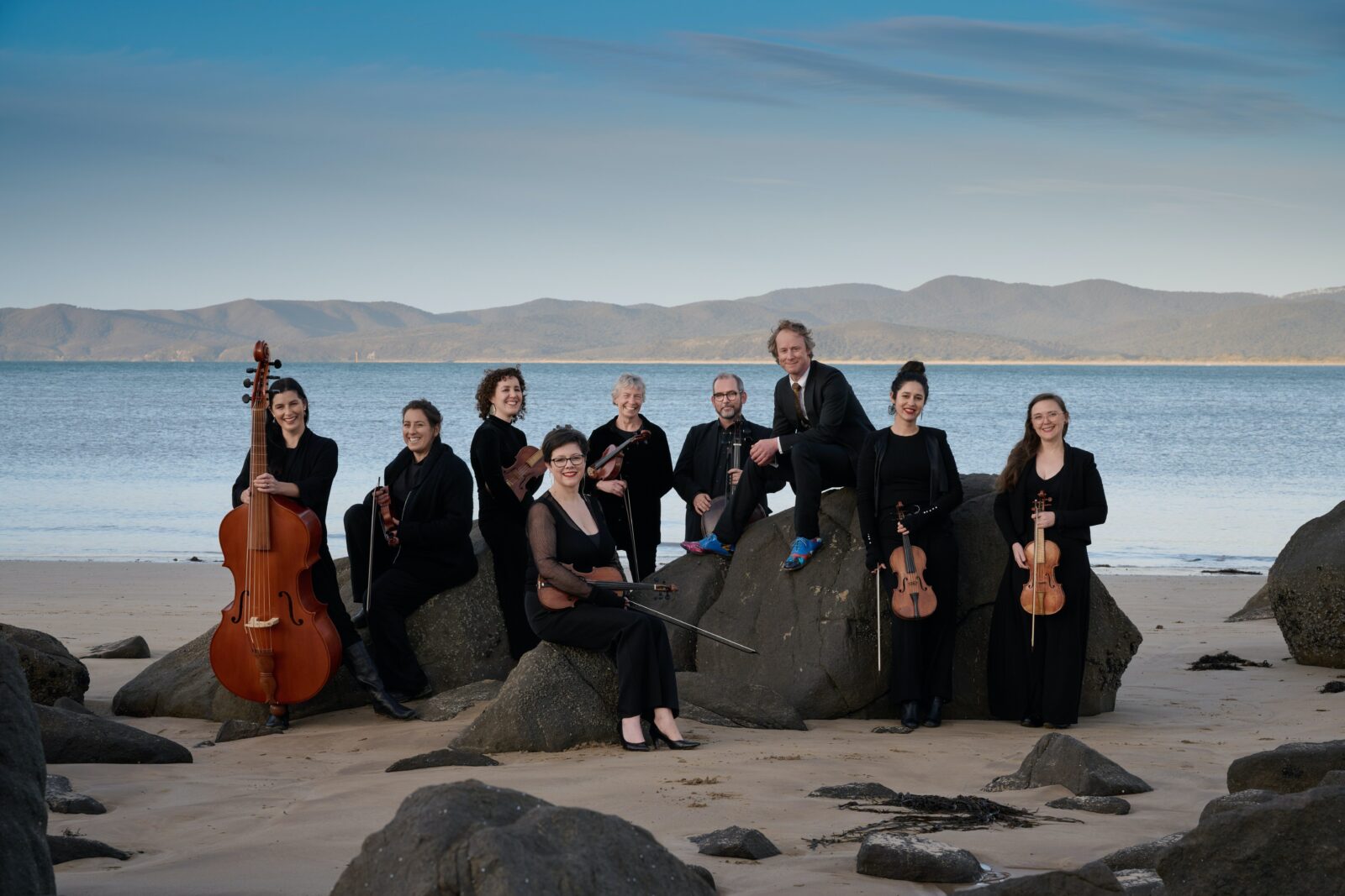 Nine smiling people gather around a group of rocks. They hold violins and other string instruments