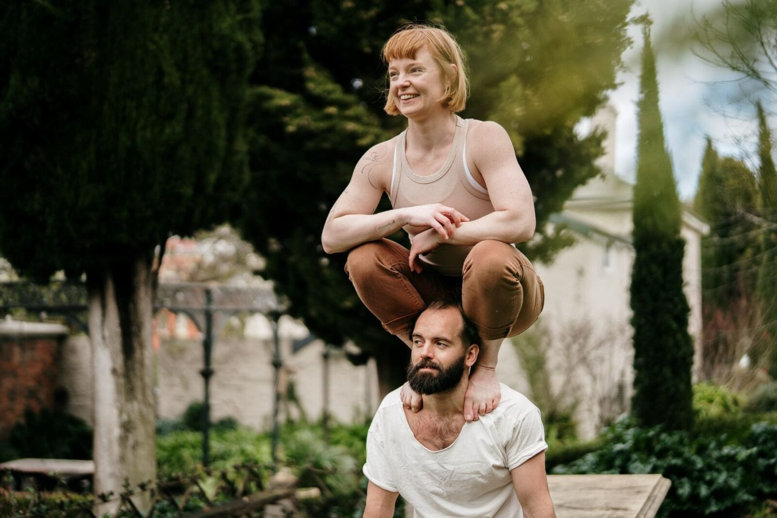 A man and a woman doing acrobatics outside. The woman is crouching on the man's shoulders.