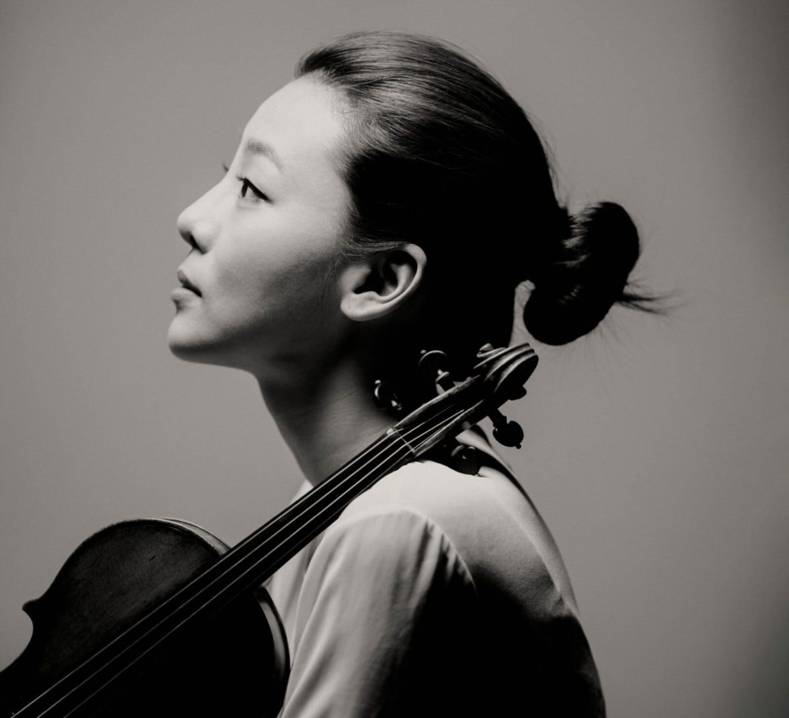Monochrome photograph of a woman, against a grey backdrop, with a violin leaning on her shoulder.