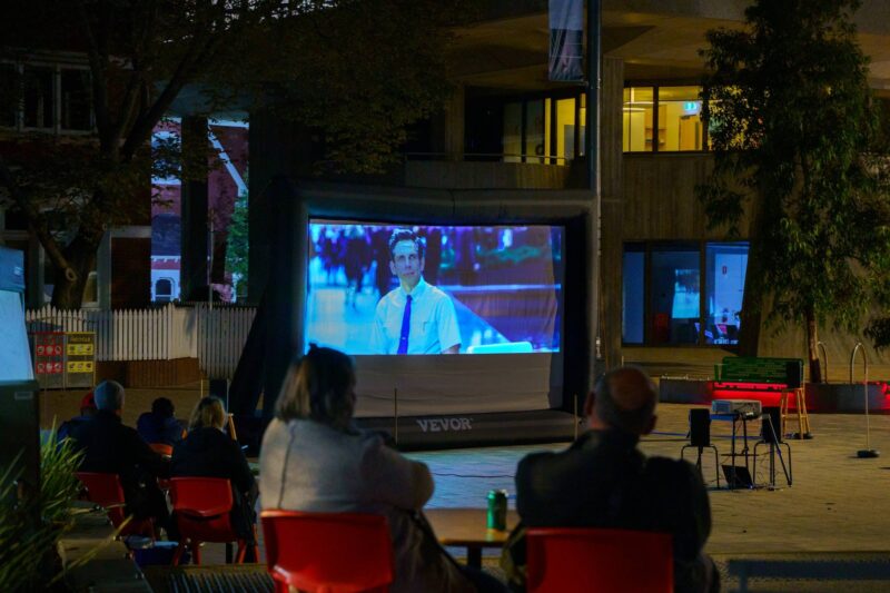 Diners watch a film on the big screen, seated at tables, outdoors.