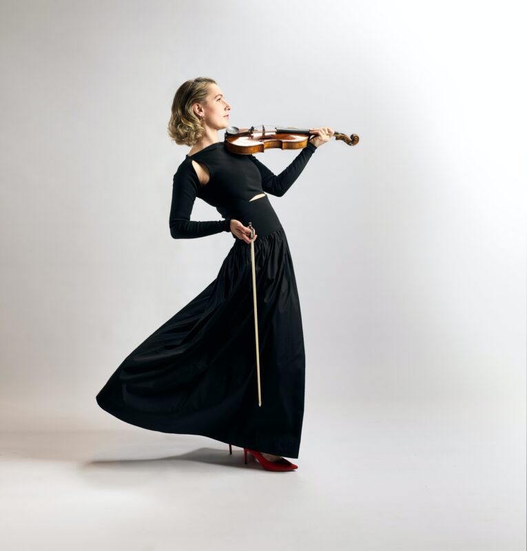 Photo of a violinist in a black dress, slightly leaning backwards, against a solid grey backdrop.