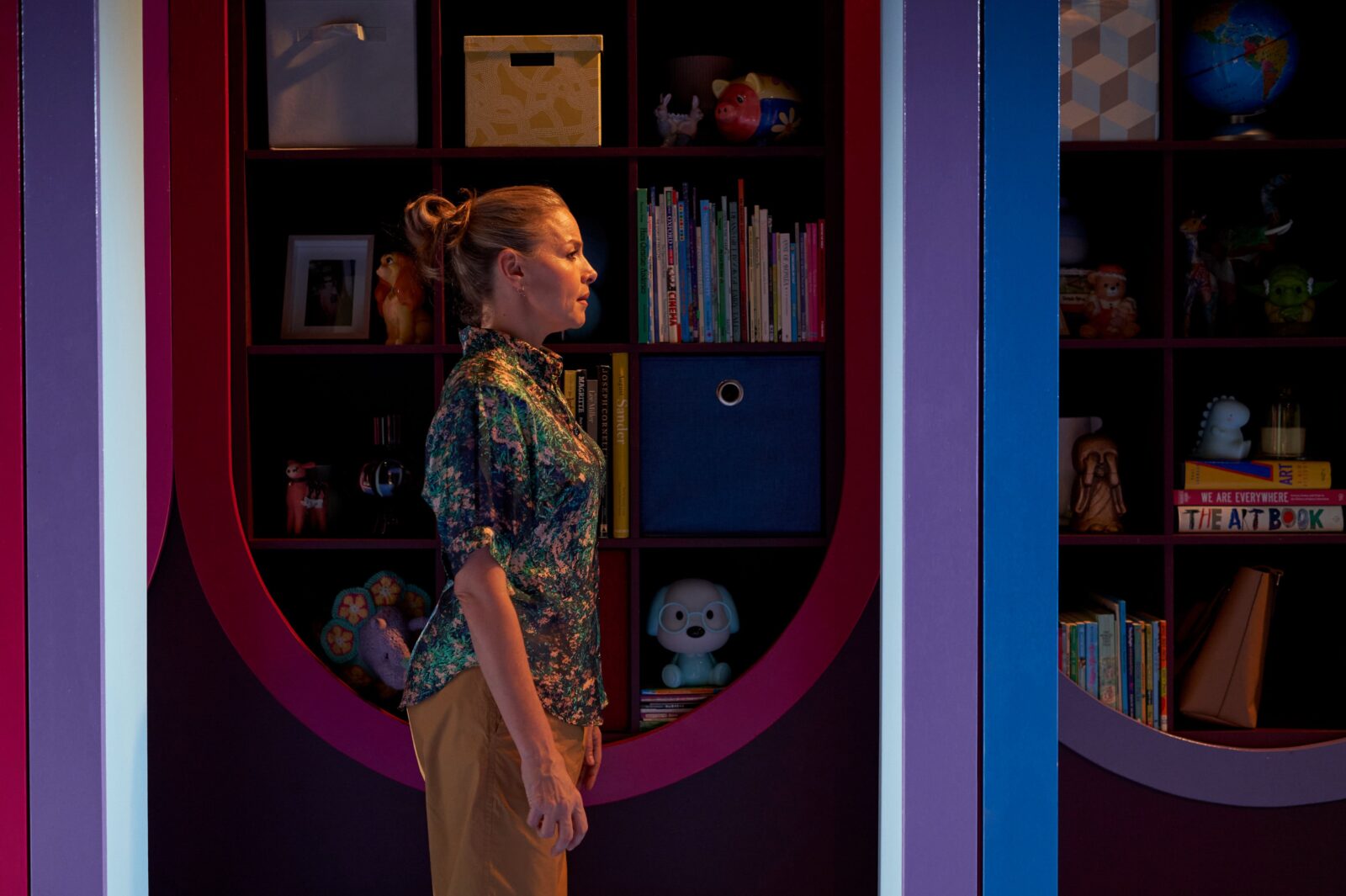 A blonde woman stands side-on to camera. Beside her a bookshelf filled with toys, books & photos