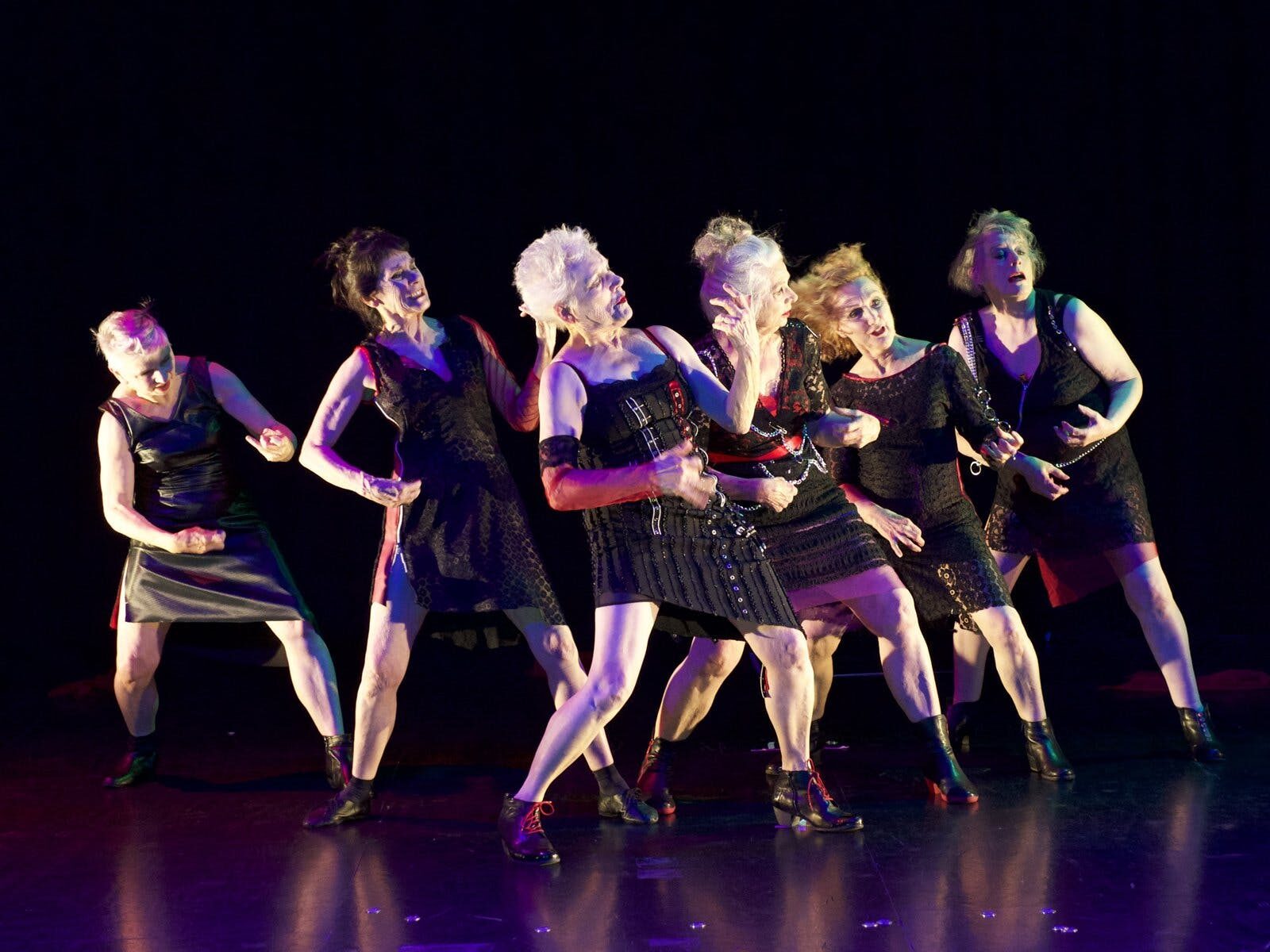 Six older women dressed in goth attire stand together, legs apart and leaning backwards