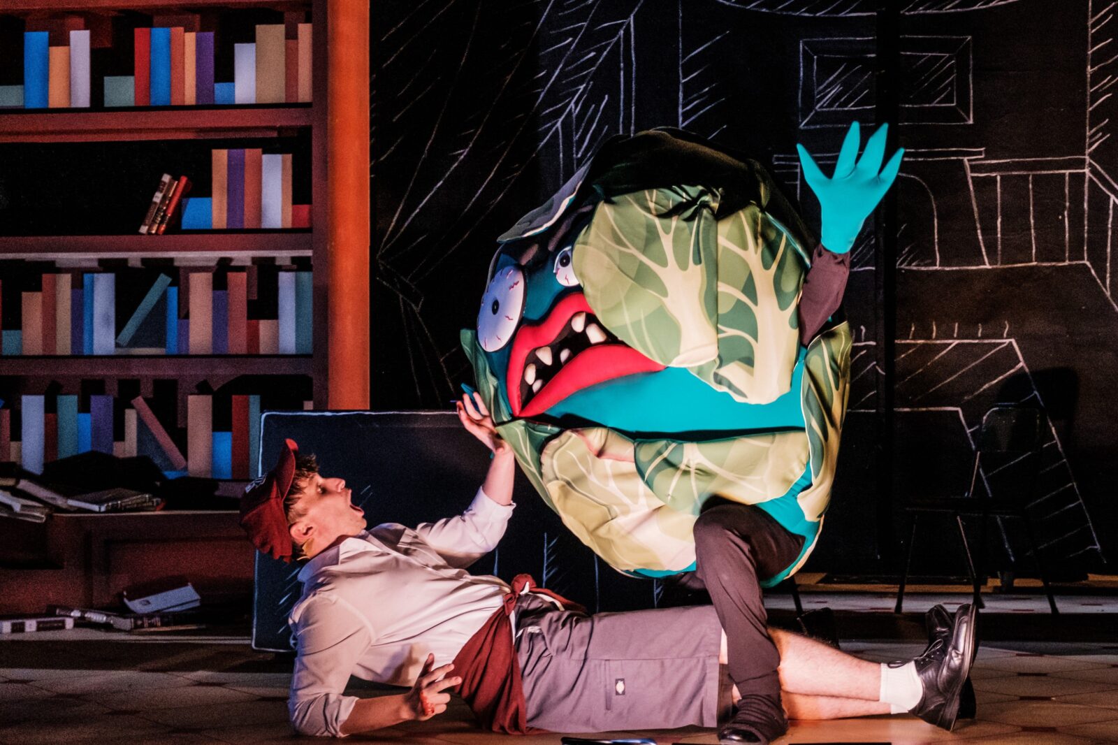 A person in a scary lettuce costume stands over a man lying on the floor dressed as a school boy.