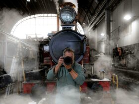 Cam Blake faces the camera with a DSLR camera held to his face. Behind him is a steam locomotive.