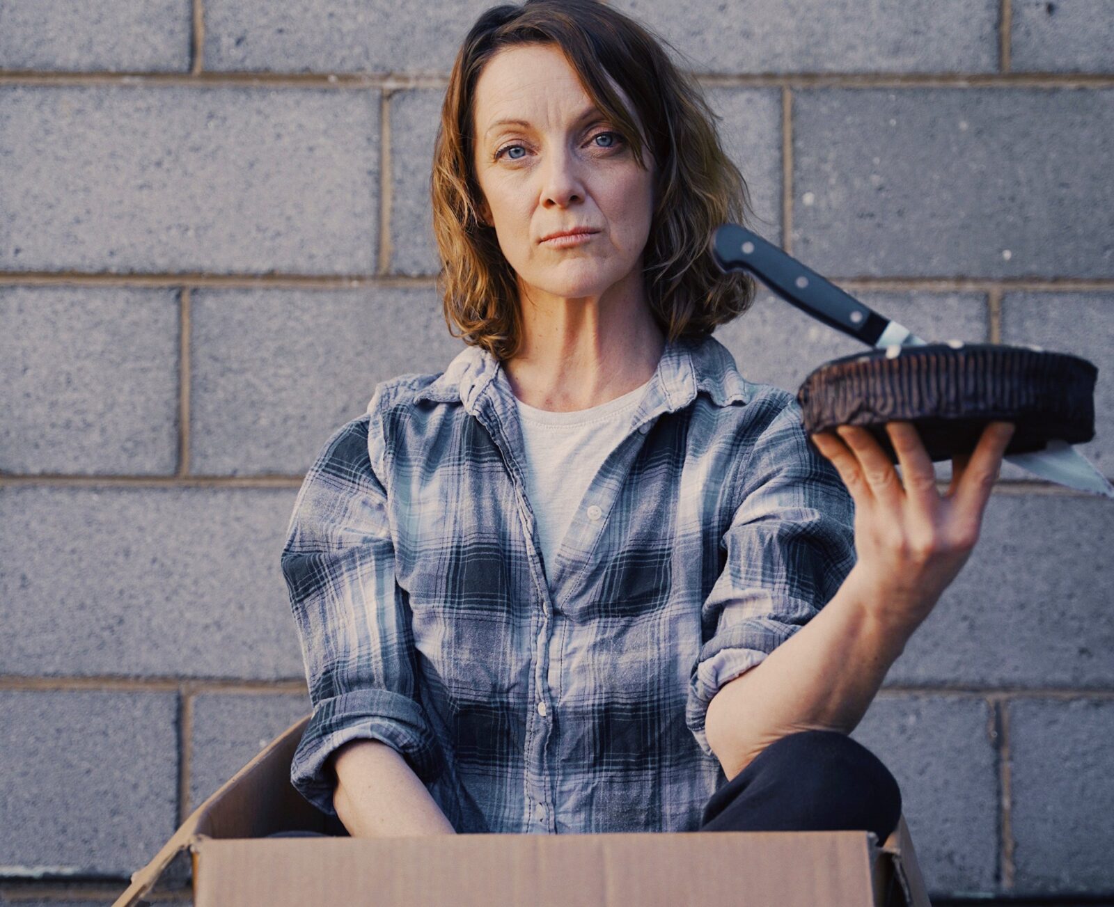 A woman in a flannelette shirt sits in a box, a woolies mudcake with a knife in her hand