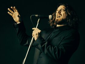 Man in all black with long hair holding a microphone and singing