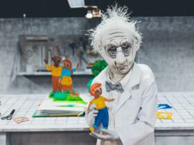 Puppet of an old man in a lab coat. He holds a small colourful child. The parents are behind waving