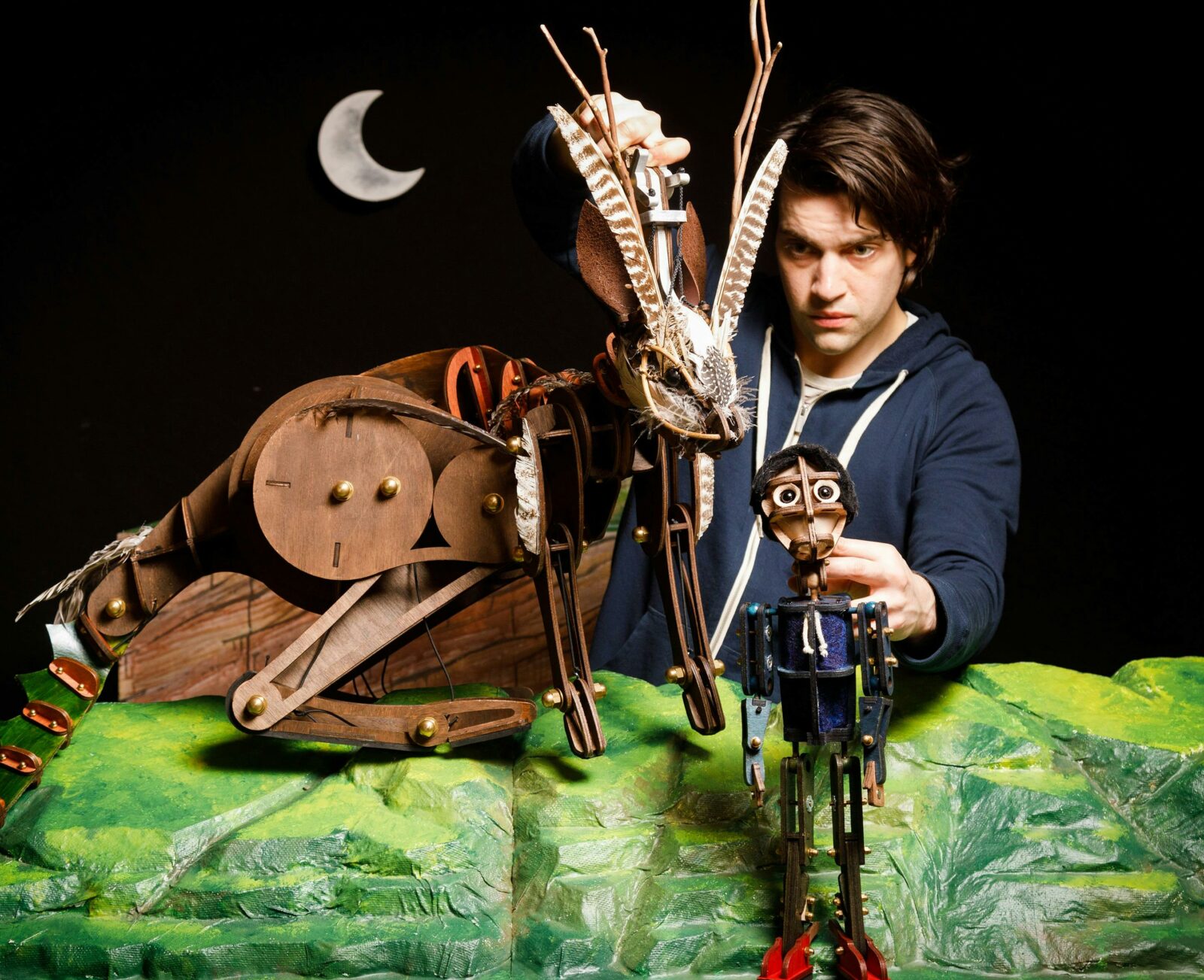 A young man holds two puppets of wood on a green landscape