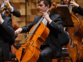 A cellist, in a black suit, playing his instrument. In the background is a double bassist.