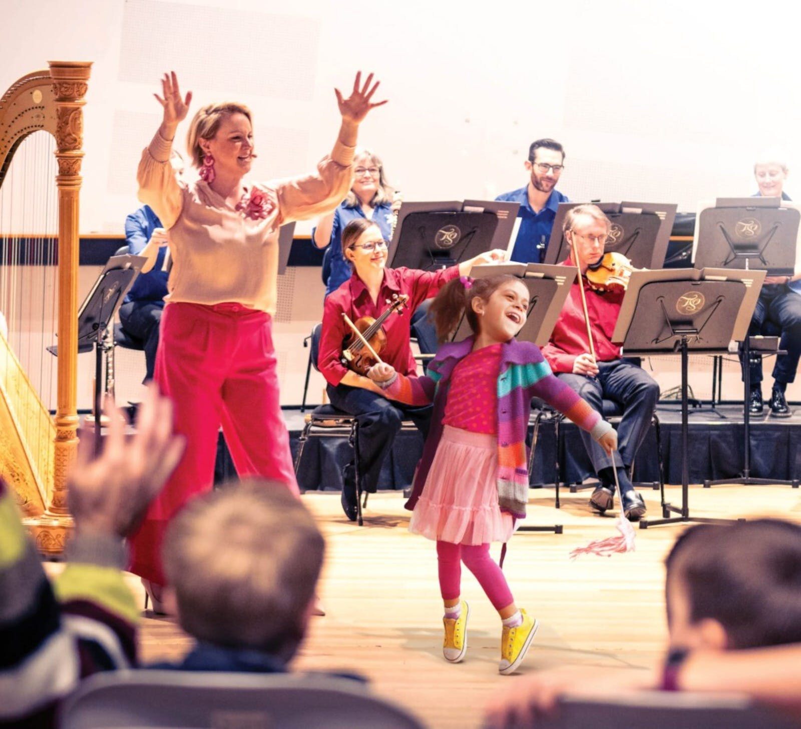 A young girl dancing in front of a musical ensemble. To the left is a harp.
