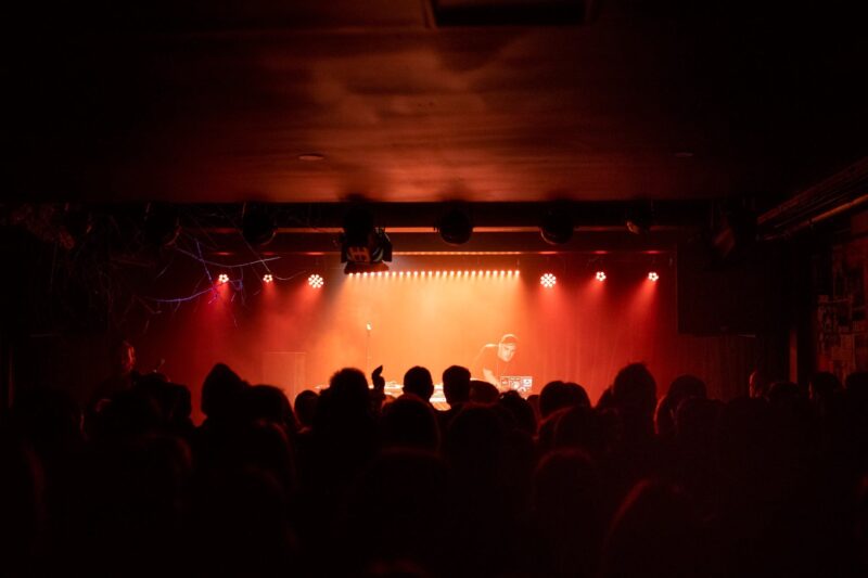 A DJ plays decks on an upraised stage with orange lighting, a crowd looks on