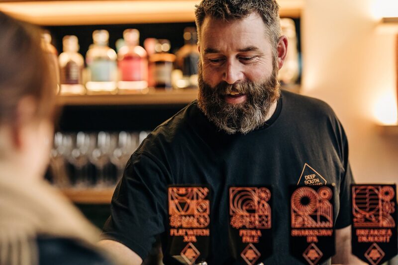 A bearded, smiling man pouring beers behind a bar