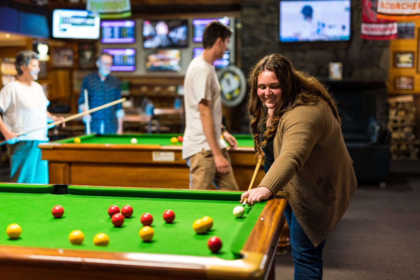 Young woman lining up a shot at one of the pool tables with a small group playing on table behind