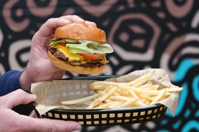 Man's hands holding double cheeseburger with lettuce and tomato and black plastic basket of fries