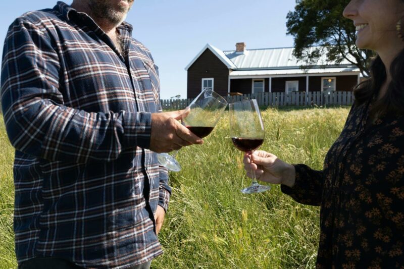 Couple toasting red wine glasses in paddock with brown weatherboard cottage in background.