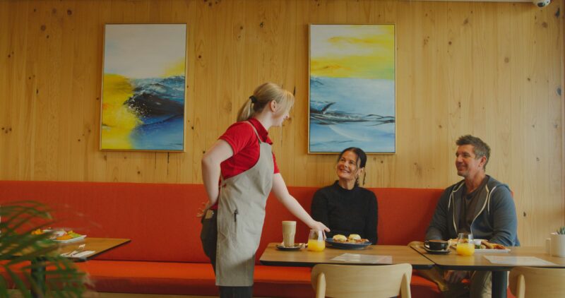 A female staff in red uniform and apron serving food for a couple of visitor sitting in the cafe