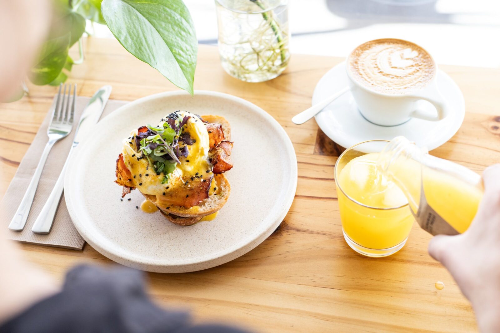 An eggs benedict served on a cream plate with a flat white and a glass of cold-pressed orange juice