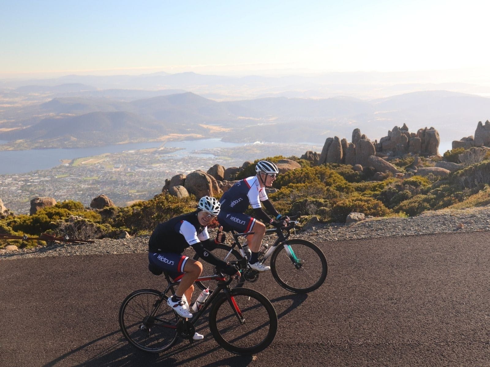 2 cyclists on bikes smiling at camera with backdrop of the city of Hobart behind them
