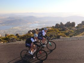 2 cyclists on bikes smiling at camera with backdrop of the city of Hobart behind them