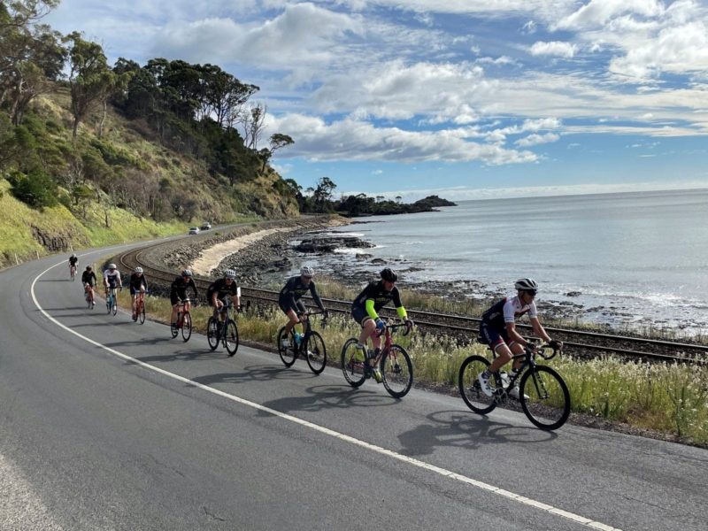 Group of cyclists riding on a coastal road with black rocks, white sand and bush setting behind.