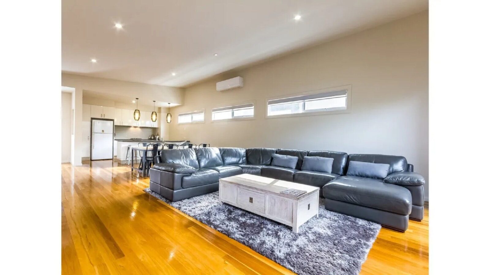 Light filled living room showing large leather corner couch and kitchen in background. Timber floor