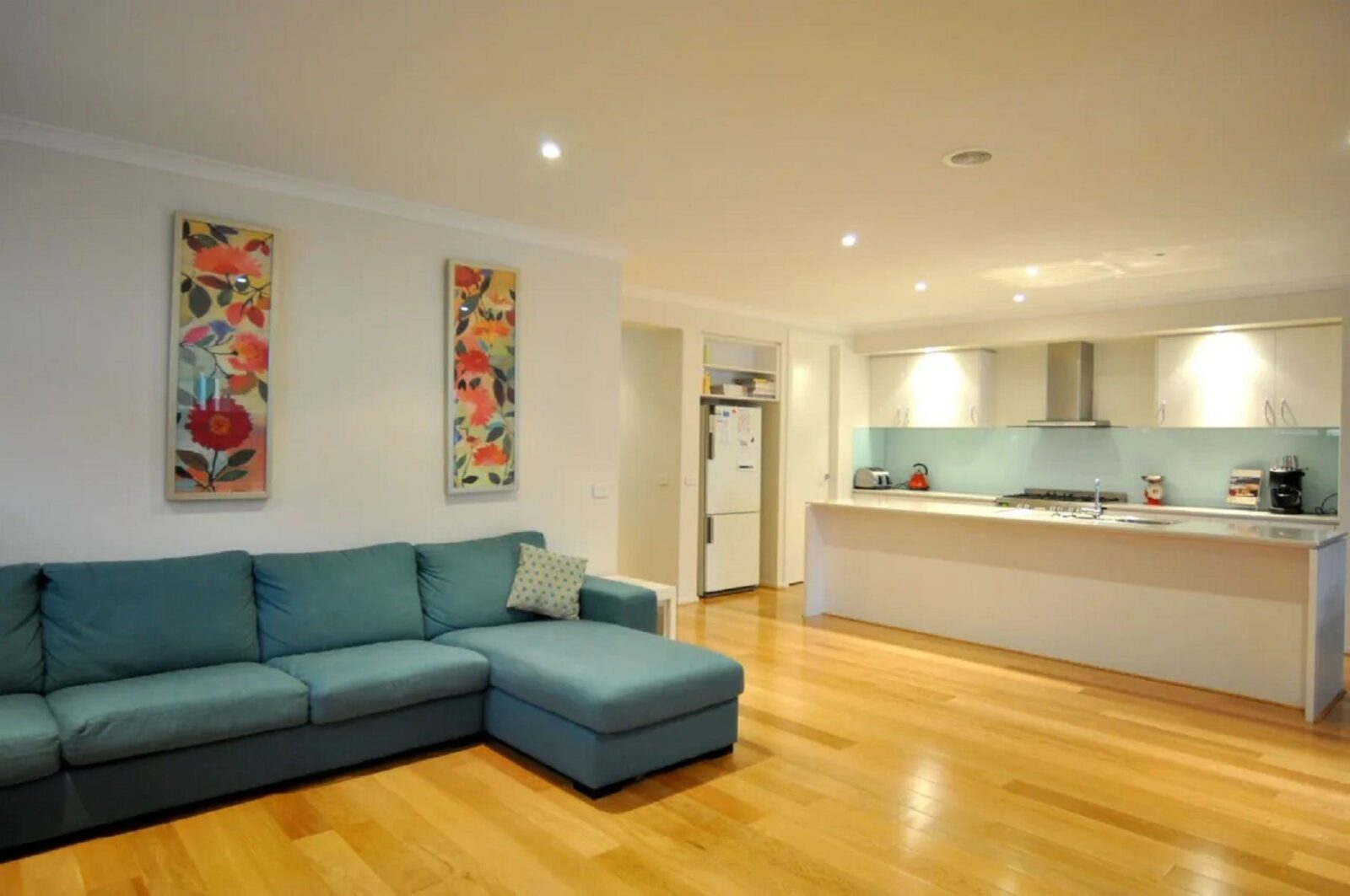 Image of main living space showing very large couch, kitchen with island bench, timber floors modern