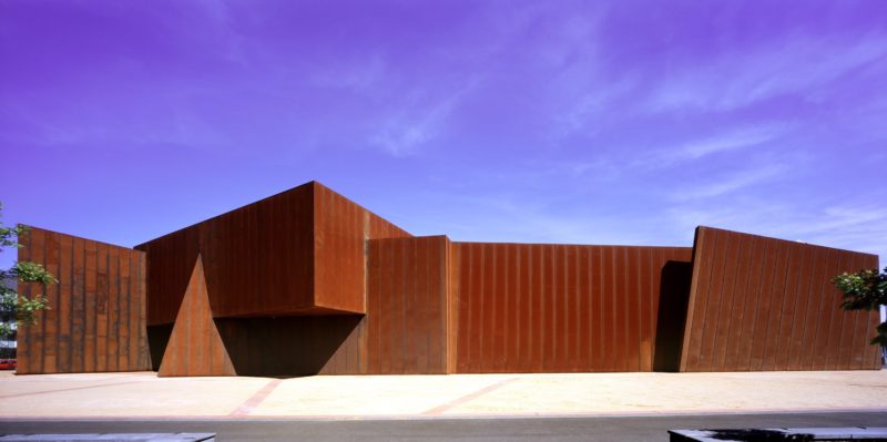 Image of a rust coloured building with sandy gravel in the foreground and a blue sky overhead