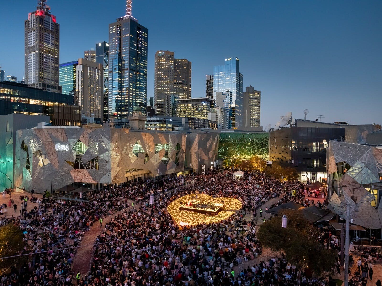Thousands gather in Fed Square around a stage, surrounded by candles in the shape of a heart.