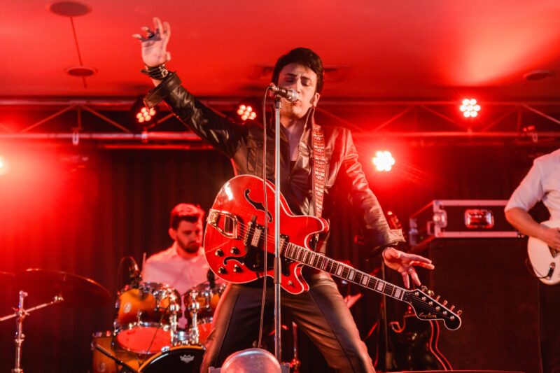 man in black leather pants and jacket with a red guitar and lighting is red