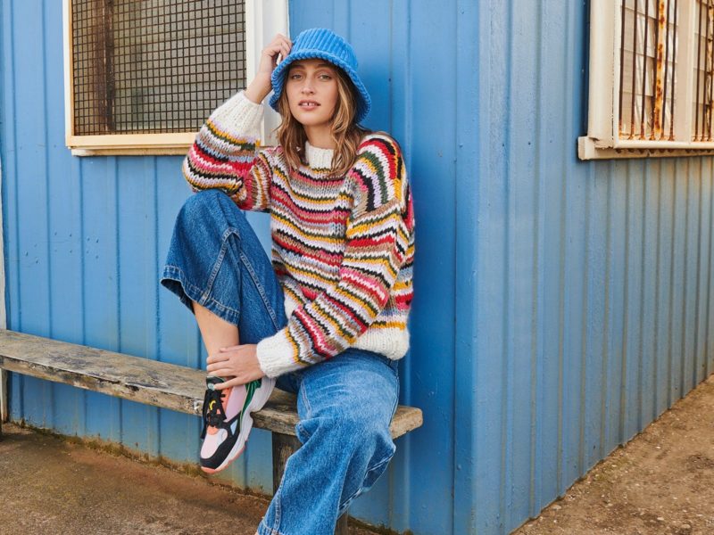 Model sits leaning against a wall with blue bucket hat, striped knit and wide leg jeans