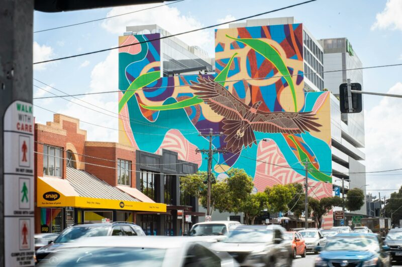 A large Indigenous mural featuring Bunjil on a building facade with cars in foreground