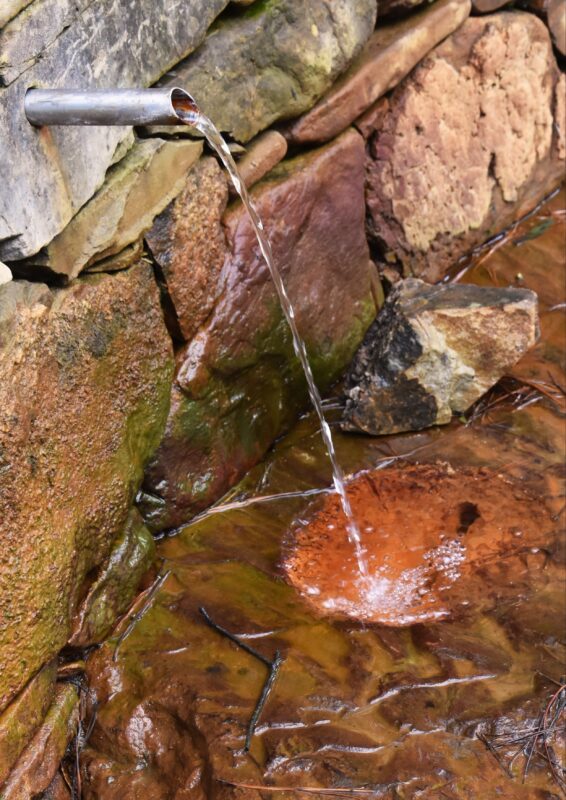 Water from the mineral spring flows from a pipe that comes out of a stone retaining wall