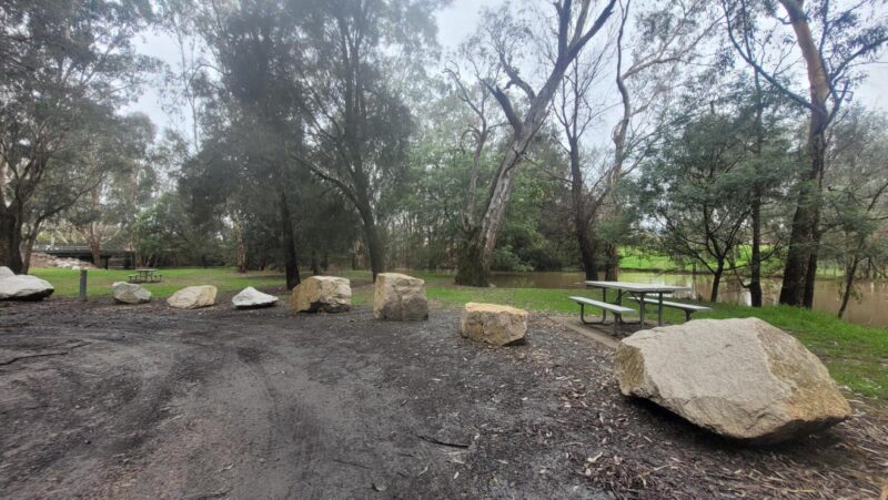 Large Rocks on side of dirt road, picnic tables, lawn, gum trees, King River, bridge in the distance