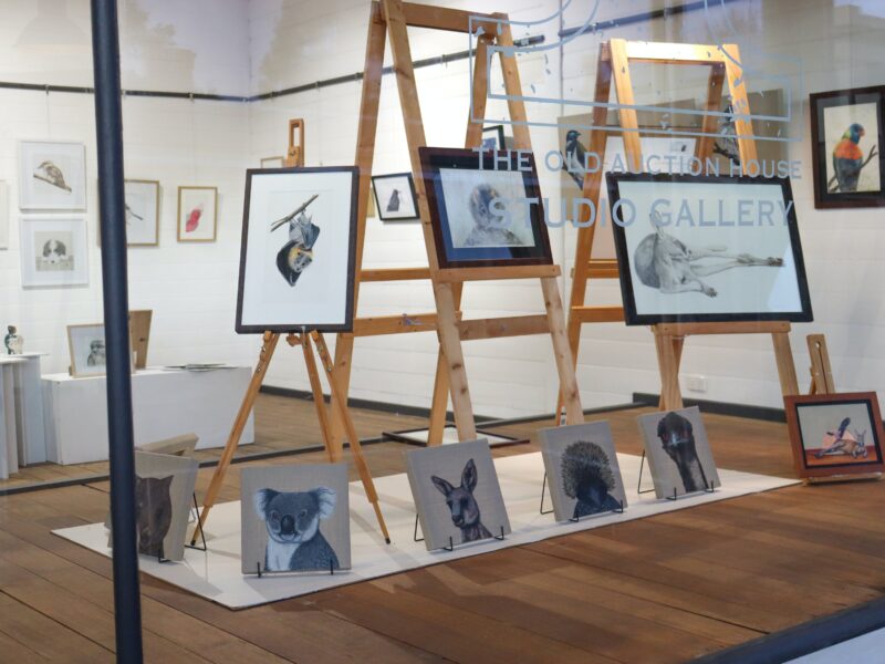 Framed artworks on easels with unframed canvases in a line in a gallery filled with art in the back