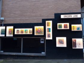 Photo of outdoor gallery with nine display boxes on a wall, lit with the an exhibition on display.