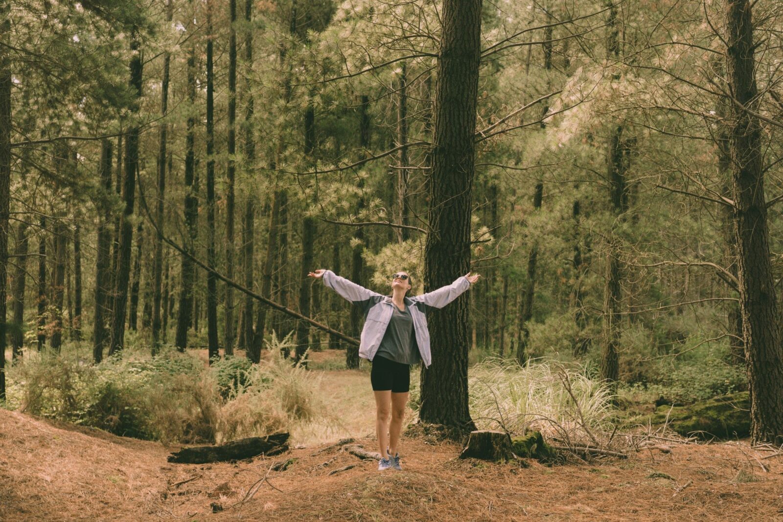 A woman in black shorts and a green shirt stands with her arms stretched out, surrounded by trees