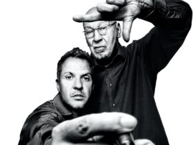 Black and white photo of a man taking a self portrait with another man framing the shot with hand