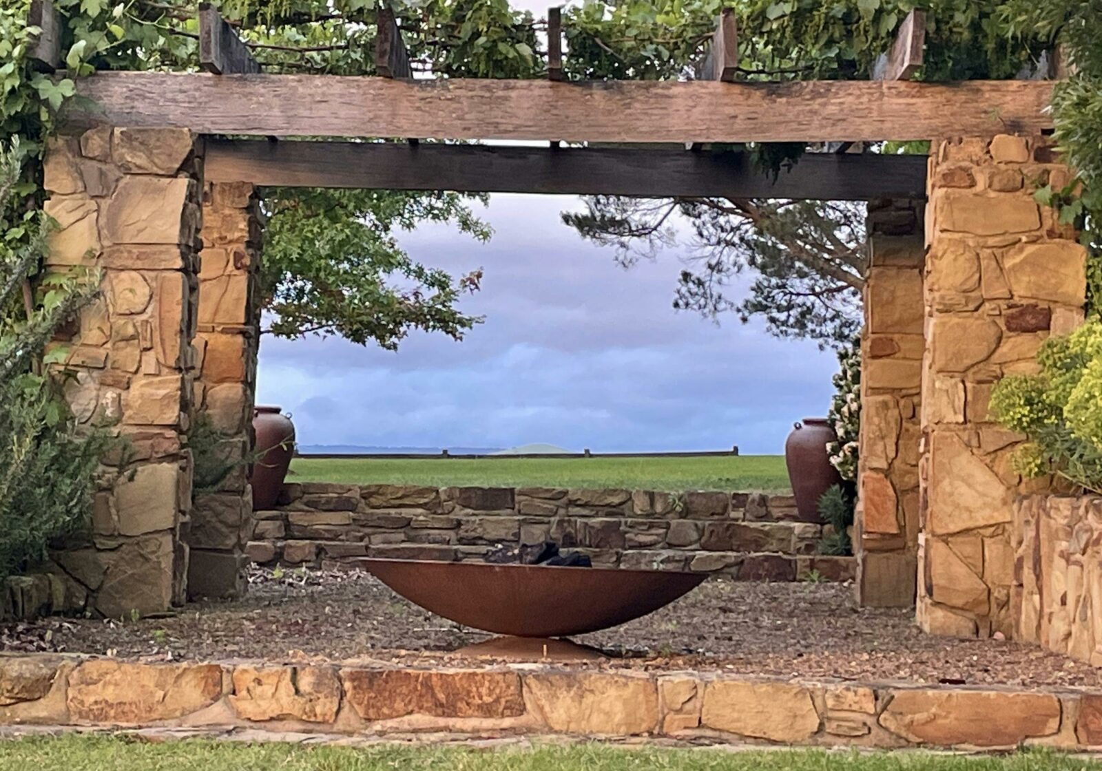 A stone and wooden arbour frames a green paddock. A fire pit sits at the front.