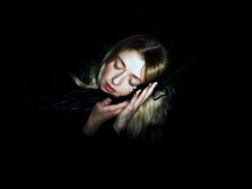 Girl with blonde hair with eyes closed leaning against black material against a black background.