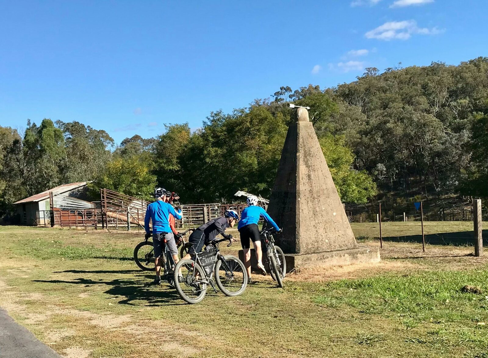 Three riders inspecting tall concrete pyramid shaped monument on roadside grassland