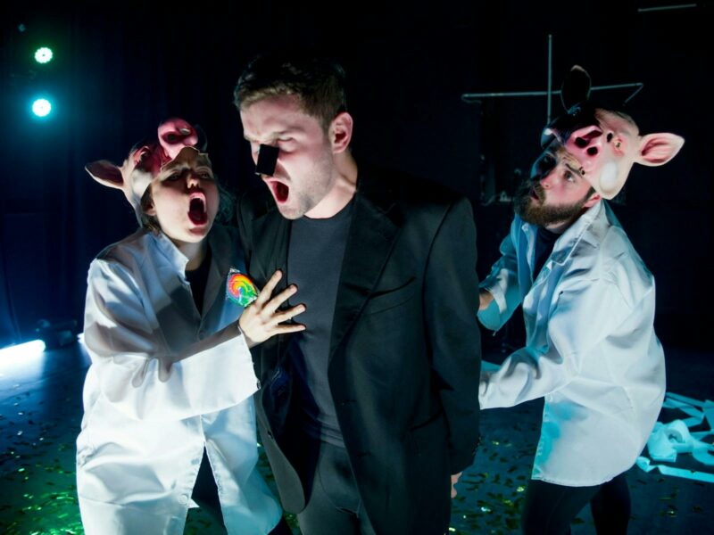 three theatre performers, two wearing pig masks and lab coats