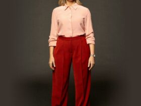 A woman with blonde hair wearing a pink shirt and red pants on a grey background.