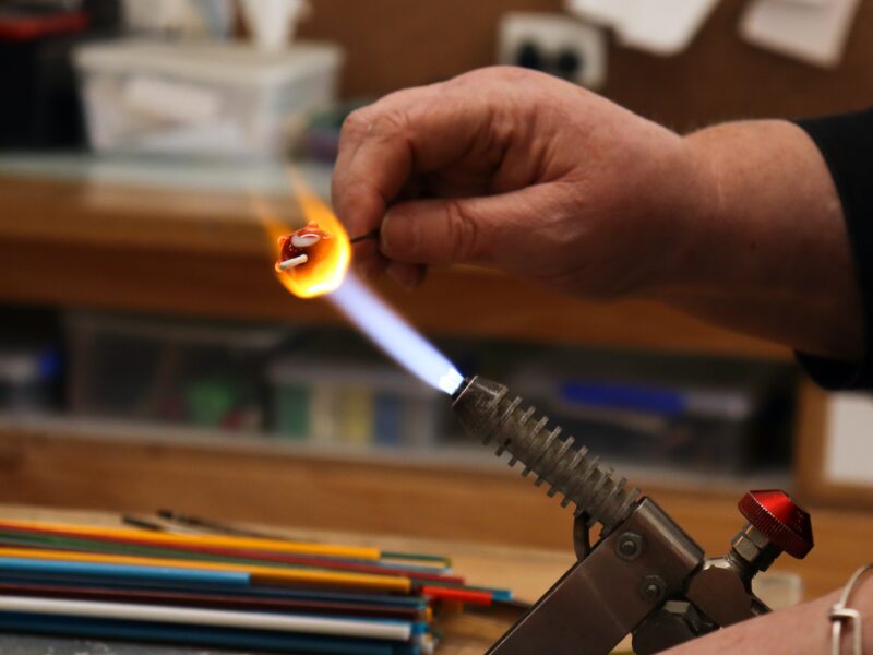 A molten glass bead glowing yellow and red being held in the blue flame of a burner by a student