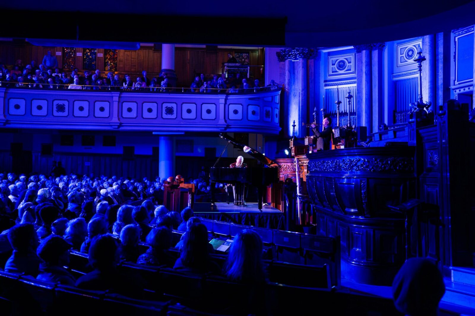 Man sitting at a grand piano on the synagogue stage, blue lighting and a captive audience