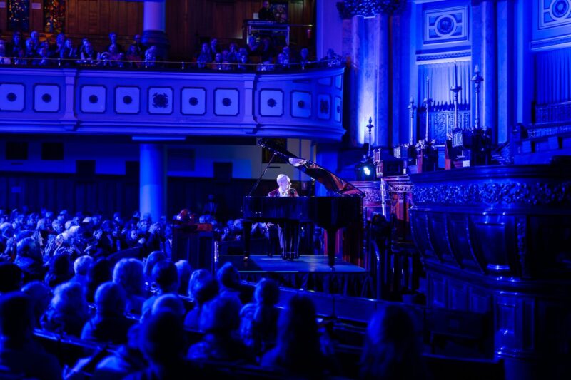 Man sitting at a grand piano on the synagogue stage, blue lighting and a captive audience