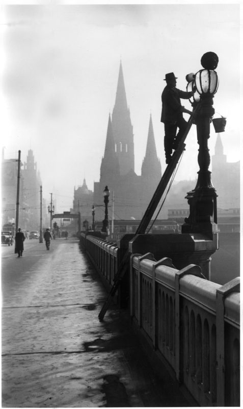 Black and while photo- a man wears a suit and hat has climbed up a ladder and is cleaning a lamppost