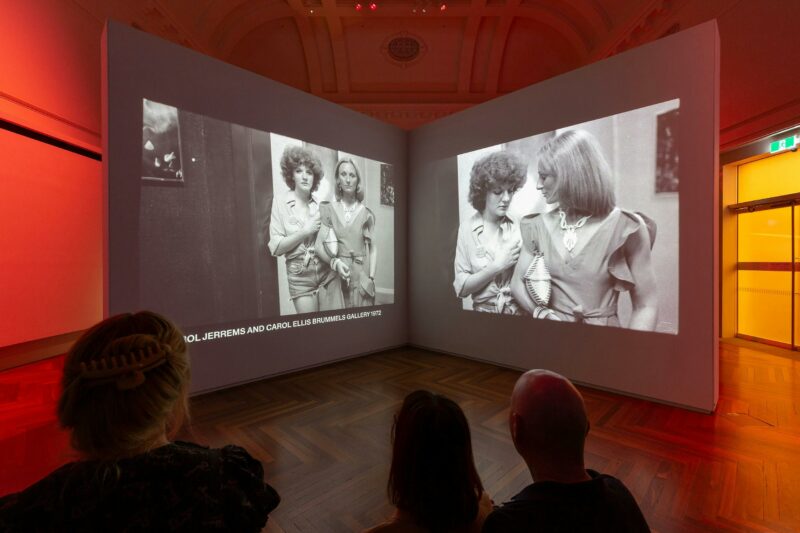 In the background are 2 screens showing black and white pictures. Heads of people in the foreground