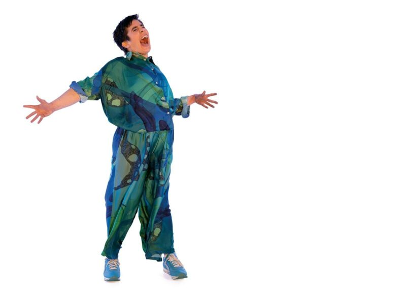 An actor wearing a blue and green jump suit with an excited expression